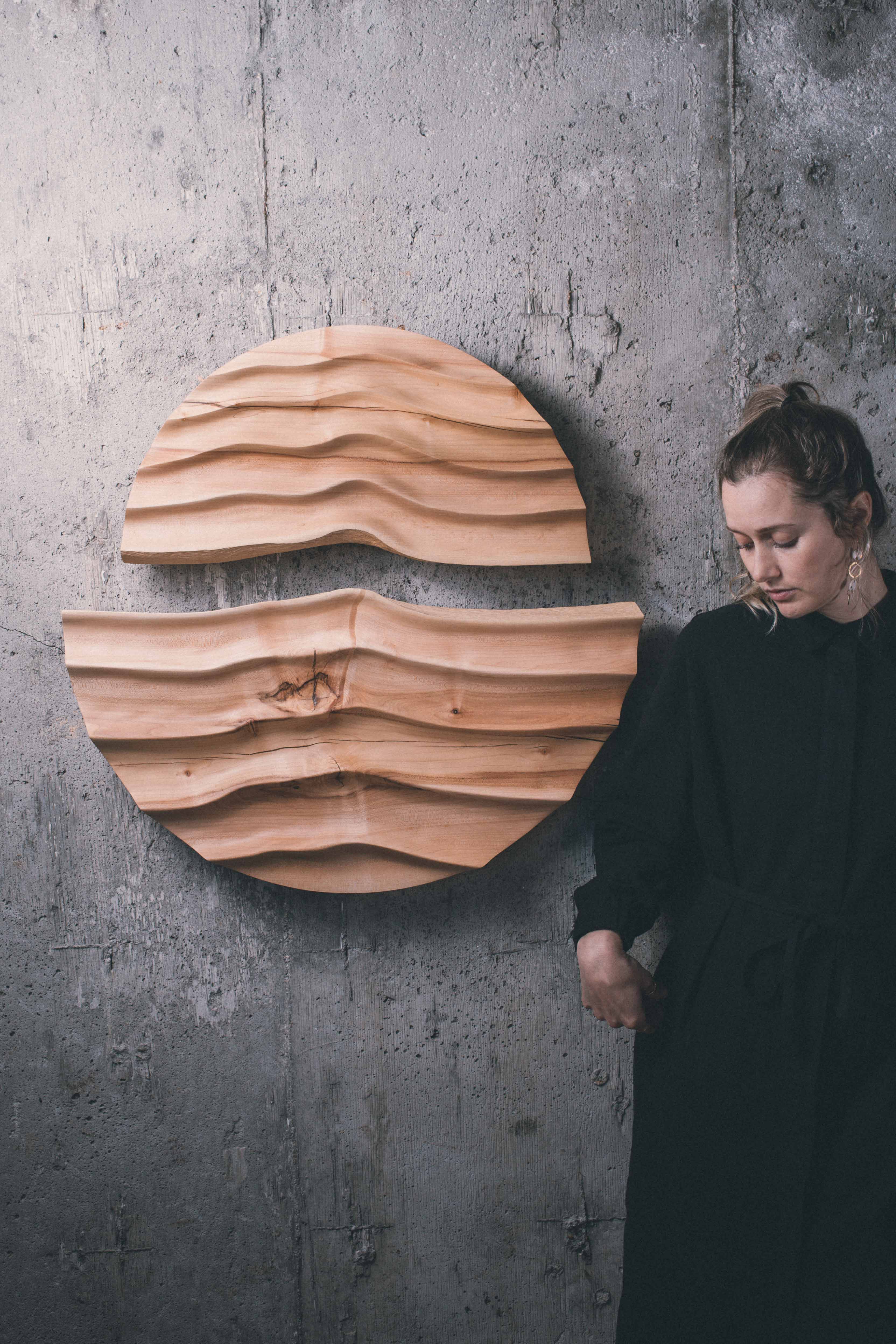 A wooden two-piece wall design hand-sculpted with a wave effect against a minimal concrete wall. To the right, artist and wood turner Jackie Chapman stands next to her work.